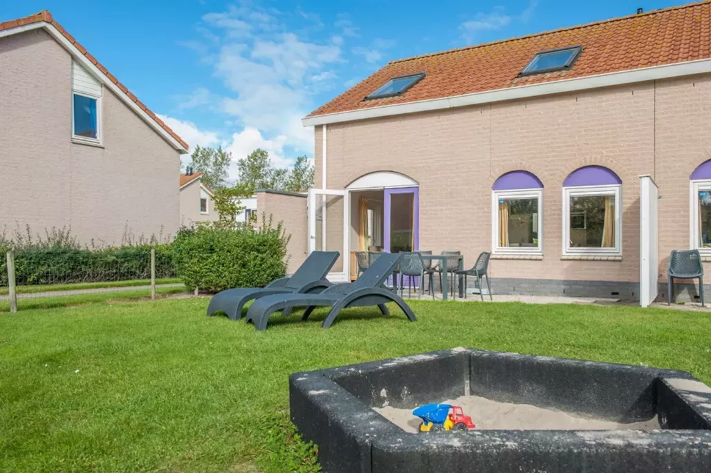 Vakantiehuisje met twee ligstoelen op het gras en zandbak met kinderspeelgoed op zonnige dag