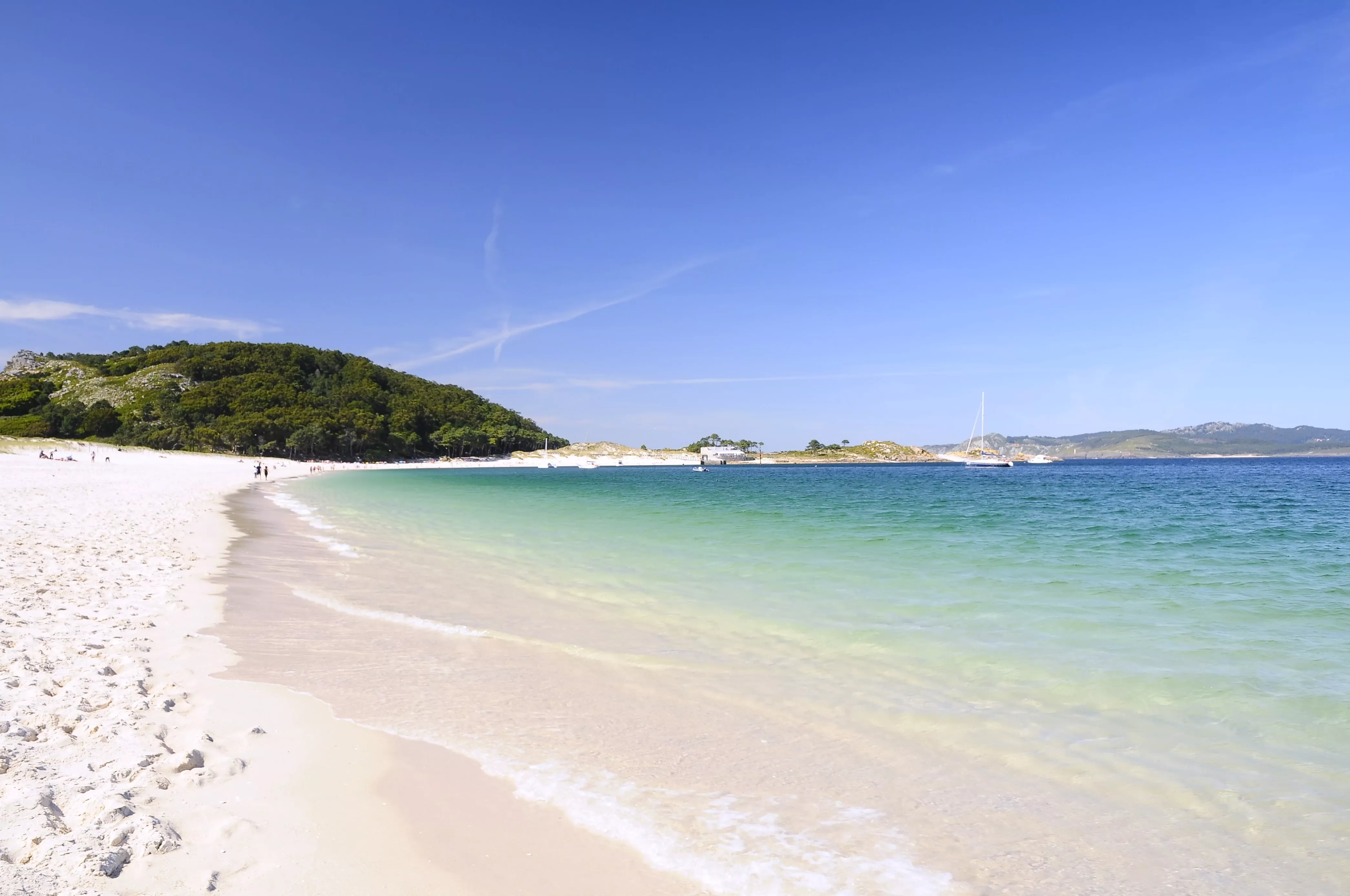 Wit zandstrand en turquoise zee op de Islas Cíes onder een strakblauwe hemel, omringd door groene heuvels.